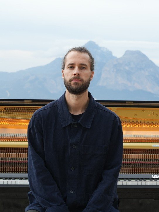 Arian M&uuml;ller seated at his piano with mountains behind